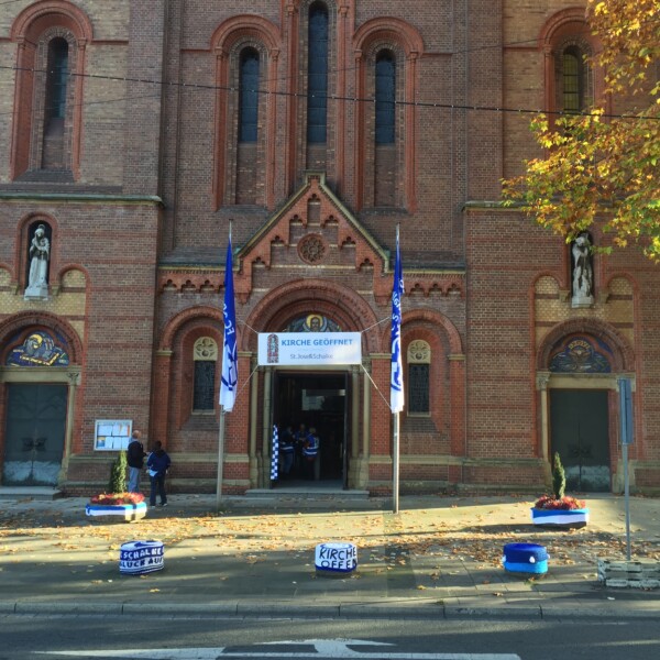 Vor dem Eingang der St.-Josephs-Kirche hängen Fahnen des FC Schalke 04. Auch die Blumenkästen und Verkehrspoller vor der Kirche sind in den Schalker Vereinsfarben geschmückt.