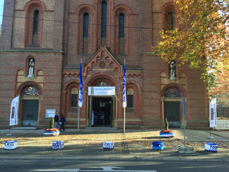 Vor dem Eingang der St.-Josephs-Kirche hängen Fahnen des FC Schalke 04. Auch die Blumenkästen und Verkehrspoller vor der Kirche sind in den Schalker Vereinsfarben geschmückt.