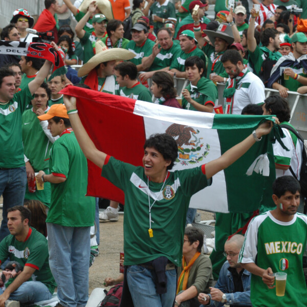 Ein junger Mann in einem Nationaltrikot von Mexiko hält mit ausgebreiteten Armen eine Mexiko-Flagge über seinen Kopf.  Hinter ihm ist eine Menschenmenge zu sehen. Die meisten tragen ebenfalls grüne Trikots von Mexiko.