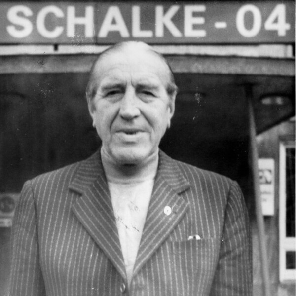 Ein alter Ernst Kuzorra steht vor unter dem Schriftzug "Schalke 04" vor dem Vereinslokal "Bosch" in Schalke und guckt in die Kamera.