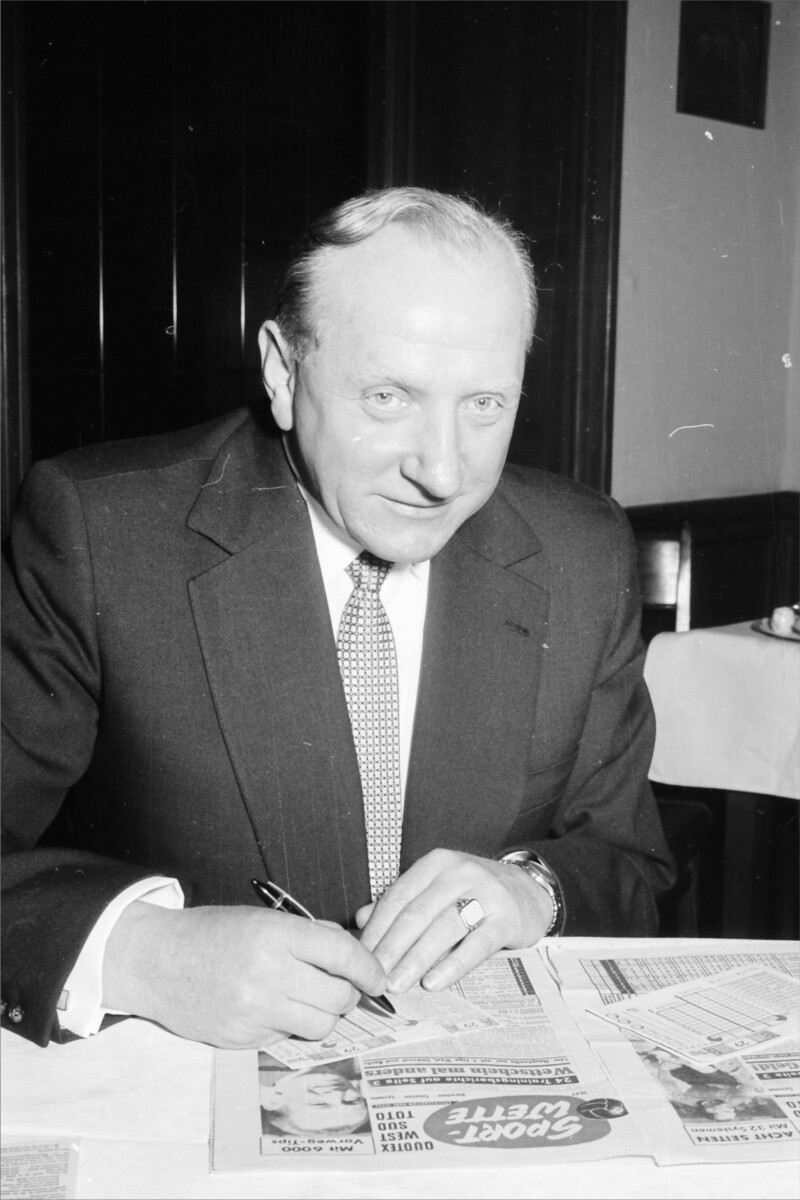 Fritz Szepan sitzt in einem Anzug und Krawatte an einem Tisch und füllt einen Wettschein aus. Unter dem Wettschein liegt eine aufgeschlagene Zeitung.