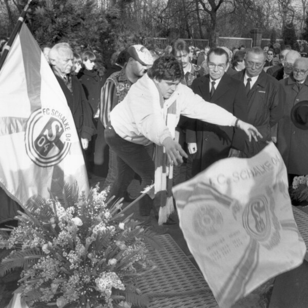 Eine große Trauergemeinde steht bei der Beisetzung auf dem Friedhof Rosenhügel am offenen Grab von Ernst Kuzorra. Ein junger Mann lässt eine Fahne vom FC Schalke 04 in das Grab fallen.