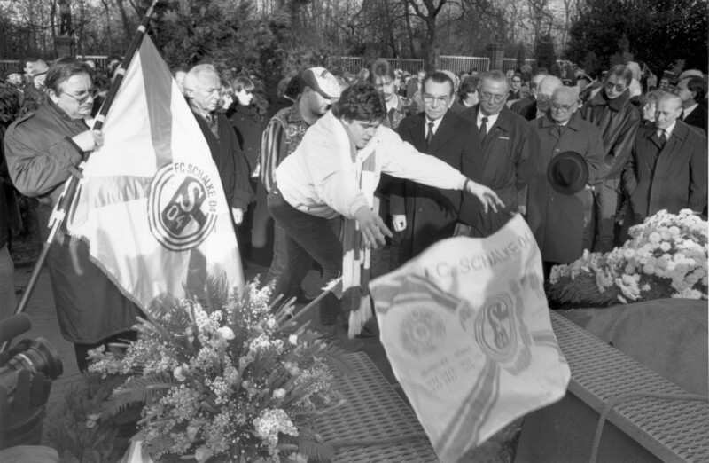 Eine große Trauergemeinde steht bei der Beisetzung auf dem Friedhof Rosenhügel am offenen Grab von Ernst Kuzorra. Ein junger Mann lässt eine Fahne vom FC Schalke 04 in das Grab fallen.