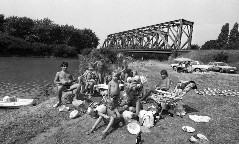 Zwei Familien sitzen mit ihren Kindern in Schwimmkleidung am Ufer des Rhein-Herne-Kanals um einen Campingtisch.