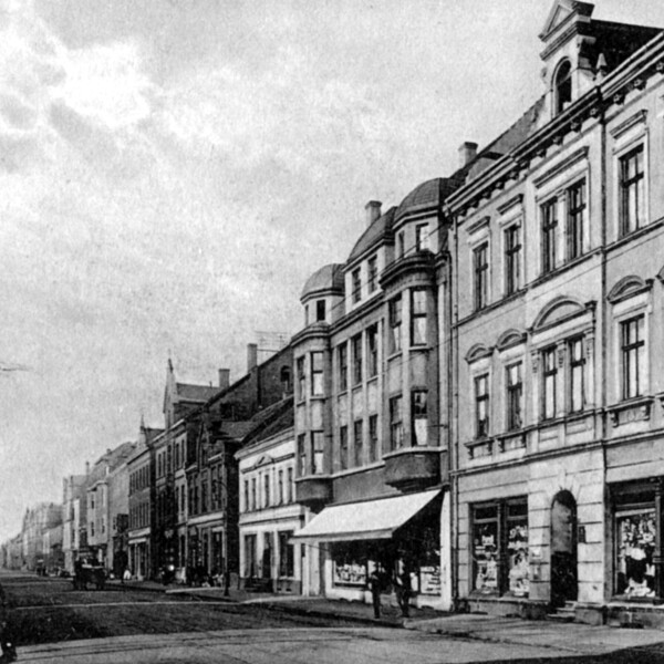 Auf der rechten Seite der nicht befestigten Straße stehen große Gründerzeithäuser mit Ladenlokalen im Erdgeschoß. Auf der linken Seite stehen einfache, eingeschoßige Häuser.