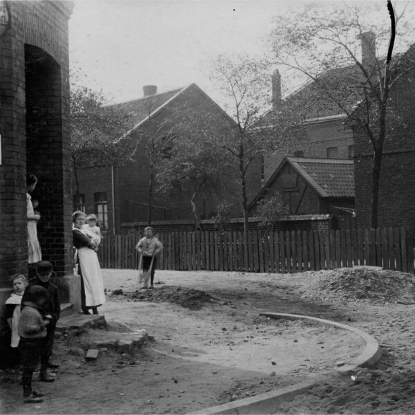 Eine Hausfrau mit einem Kleinkind auf dem Arm und ein paar Kinder an einer Straßenecke in Schalke um 1900. Die Straße und der Bürgersteig sind nicht befestigt. Hinter ihnen ist ein mit einfachen Häusern dicht bebautes Wohnquartier zu erkennen.