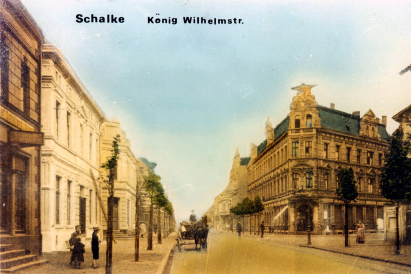 Eine Ansichtskarte der König-Wilhelm-Straße in Schalke um die Jahrhundertwende. Die Straße ist befestigt und gepflegt. Die Bürgersteige sind mit Bäumen bepflanzt.
