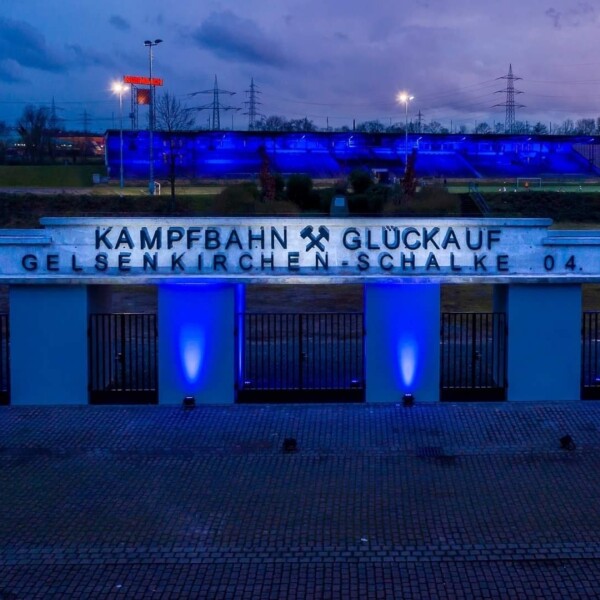 Das Eingangstor an der Kampfbahn Glückauf wird in der Dämmerung von vier Strahlern blau beleuchtet. Auch die Tribüne im Hintergrund ist in blaues Licht getaucht.