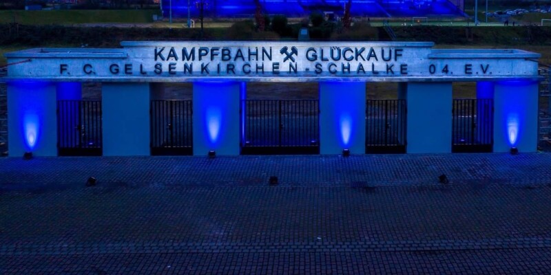 Das Eingangstor an der Kampfbahn Glückauf wird in der Dämmerung von vier Strahlern blau beleuchtet. Auch die Tribüne im Hintergrund ist in blaues Licht getaucht.
