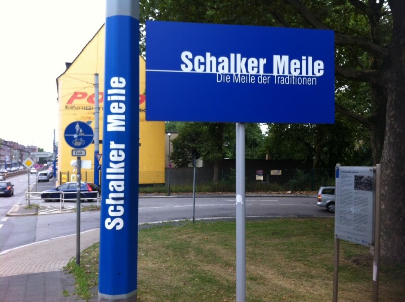 Auf einem blauen Schild steht in weißer Schrift "Schalker Meile - Die Meile der Tradition". Es ist an einer Straßenlaterne angebracht.