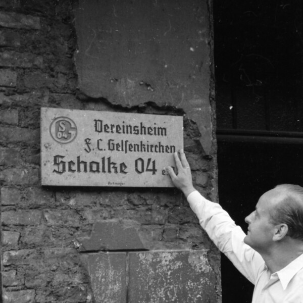 Otto Tibulsky berührt mit seiner rechten Hand ein Eingangsschild. Auf dem Schild steht "Vereinsheim FC Schalke 04". Die Fassade des Hauses zeigt Kriegsschäden: An vielen Stellen fehlt der Putz, das Mauerwerk ist sichtbar.