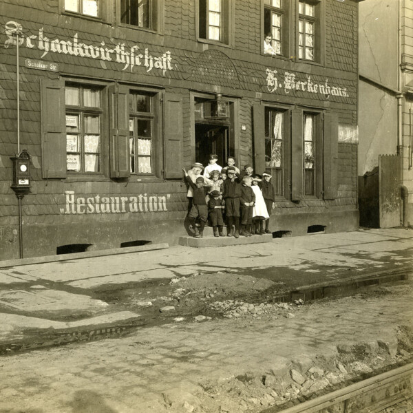 Vor dem Eingang eines Mehrfamilienhauses posiert eine Gruppe aus acht Jugendlichen und Kindern. Auf der Fassade des Hauses stehen die Worte "Schankwirtschaft" und "Restauration".