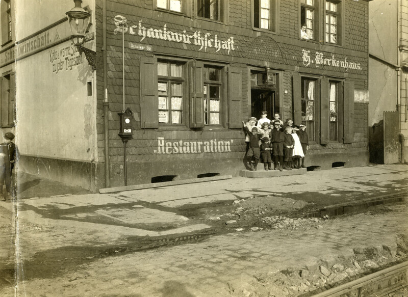 Vor dem Eingang eines Mehrfamilienhauses posiert eine Gruppe aus acht Jugendlichen und Kindern. Auf der Fassade des Hauses stehen die Worte "Schankwirtschaft" und "Restauration".