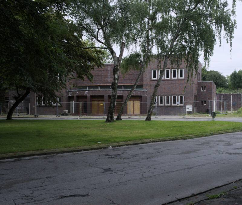 Das Gebäude vom Bahnhof Schalke-Nord liegt verlassen hinter zwei Bäumen. Die Zugänge des Haupteingangs sind mit Brettern vernagelt.