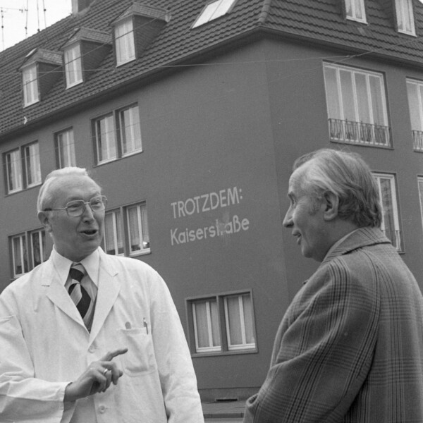 Ein älterer Mann in einem weißen Kittel unterhält sich mit einem anderen Mann. Zwischen ihnen ist an der Fassade eines Hauses im Hintergrund ist in großen weißen Buchstaben die Aufschrift "Trotzdem: Kaiserstraße" zu lesen.