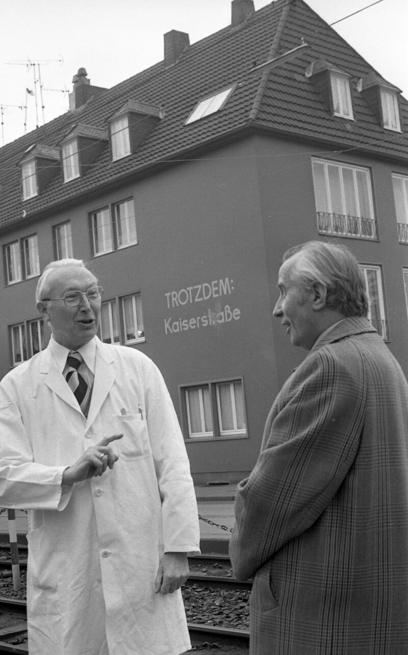 Ein älterer Mann in einem weißen Kittel unterhält sich mit einem anderen Mann. Zwischen ihnen ist an der Fassade eines Hauses im Hintergrund ist in großen weißen Buchstaben die Aufschrift "Trotzdem: Kaiserstraße" zu lesen.
