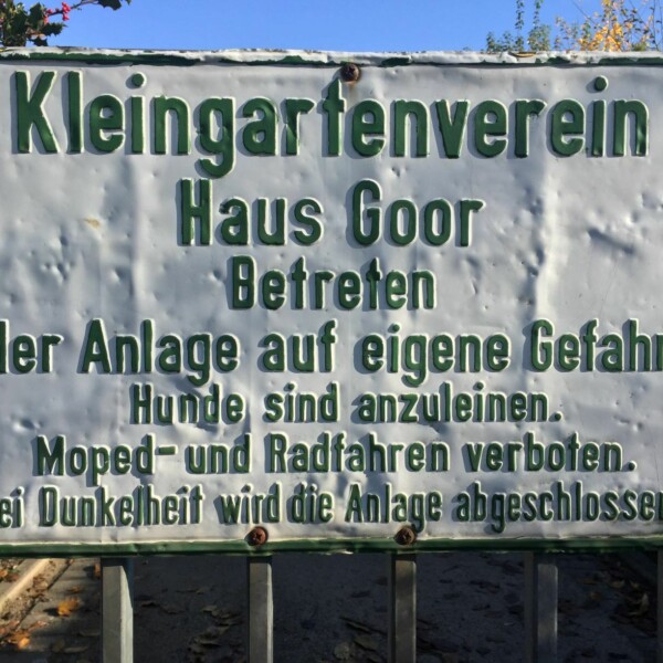 Ein weißes, zerbeultes Schild, auf dem in grüner Schrift "Kleingartenverein Haus Goor" steht.