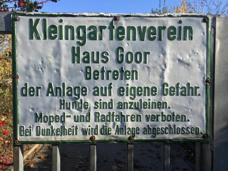 Ein weißes, zerbeultes Schild, auf dem in grüner Schrift "Kleingartenverein Haus Goor" steht.