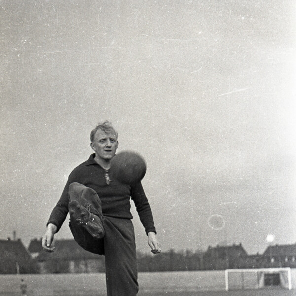 Fritz Szepan steht in Trainingsklamotten auf einem Sportplatz. Er nimmt einen Ball auf Brusthöhe mit seinem rechten Fuß an.