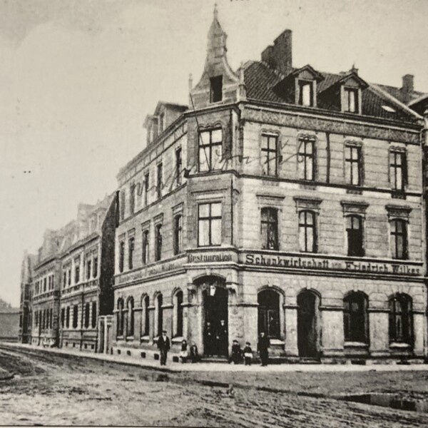 Blick in eine unbefestigte Straße. Auf der Ecke steht ein großes Haus. Über dem Erdgeschoss steht "Restauration und Schankwirtschaft Friedrich Wilkes".