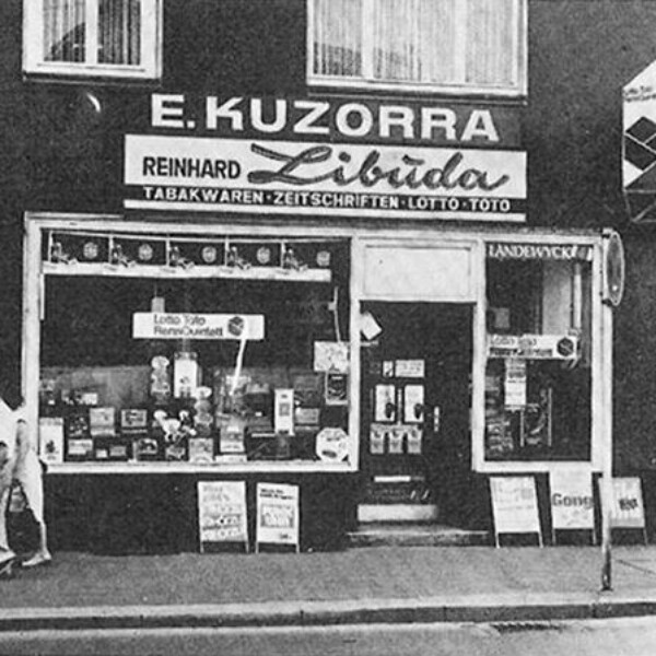 Frontansicht des Tabakladens. Über dem Eingang hängt ein Schild mit der Aufschrift "E. Kuzorra, Reinhard Libuda".