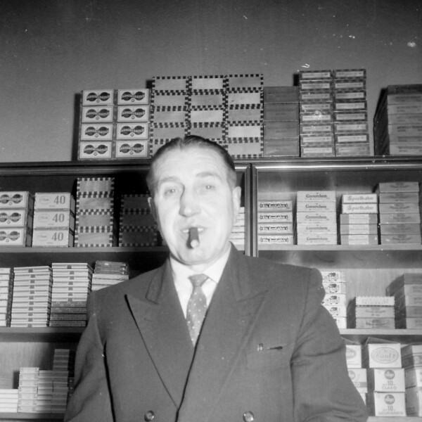 Ernst Kuzorra hält in seiner Hand eine Rolle Papier. Im Mund hält er eine Zigarre. Hinter ihm steht ein Regal mit Tabakwaren.