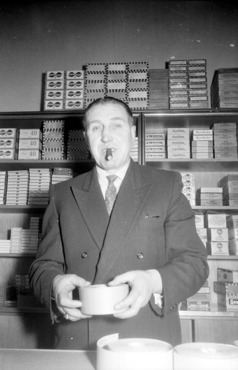 Ernst Kuzorra hält in seiner Hand eine Rolle Papier. Im Mund hält er eine Zigarre. Hinter ihm steht ein Regal mit Tabakwaren.