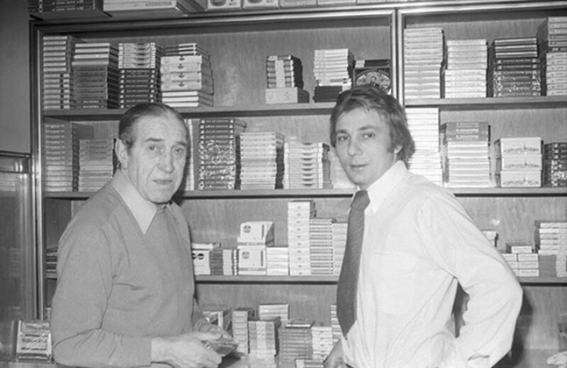 Ernst Kuzorra und Stan Libuda stehen gemeinsam vor einem Regal mit Tabakwaren und gucken in die Kamera.