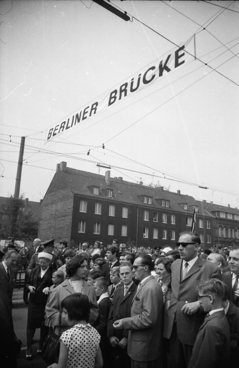Die Straße ist voll mit Menschen. Die Männer und Jungen tragen Anzüge, die Frauen festliche Kleider. Über ihnen hängt in etwa vier Metern Höhe der Schriftzug "Berliner Brücke".