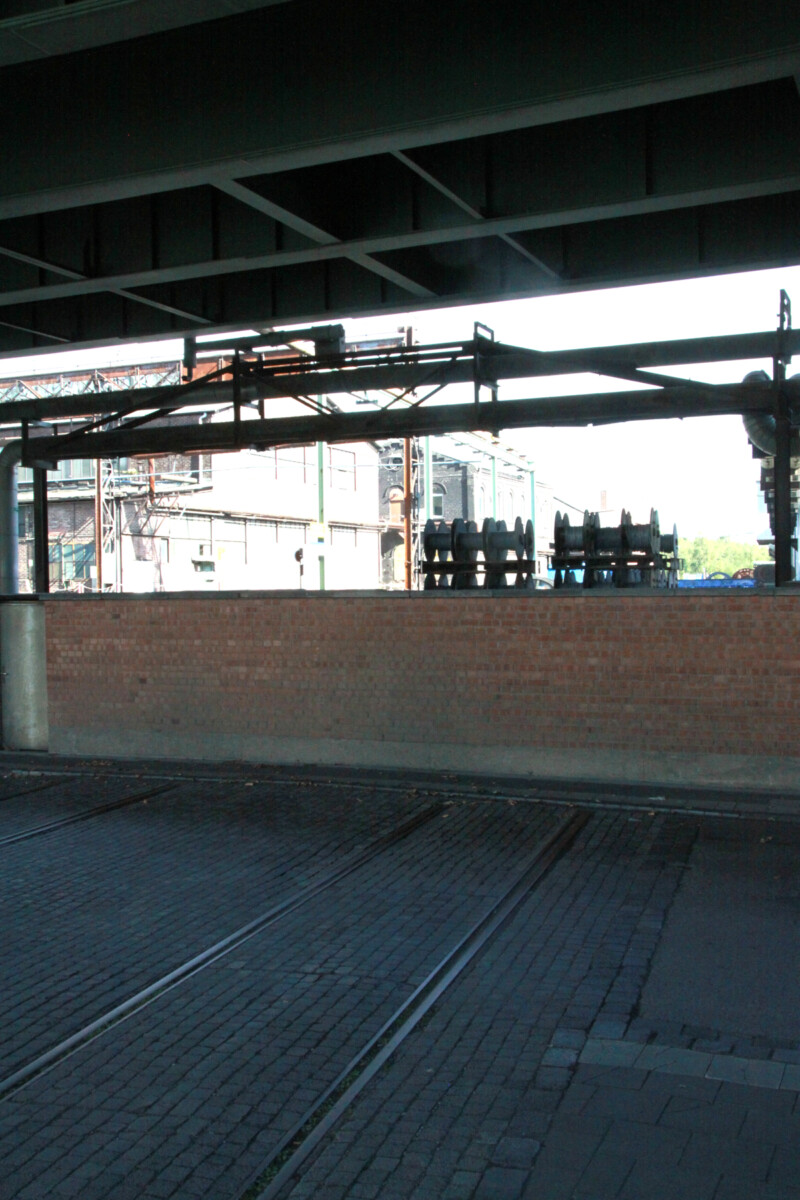 Eine in der Straße eingebettete Schiene der ehemaligen Zechenbahn der Zeche Consolidation 1/6 endet an einer Mauer am Schalker Markt. Darüber verläuft die Berliner Brücke.