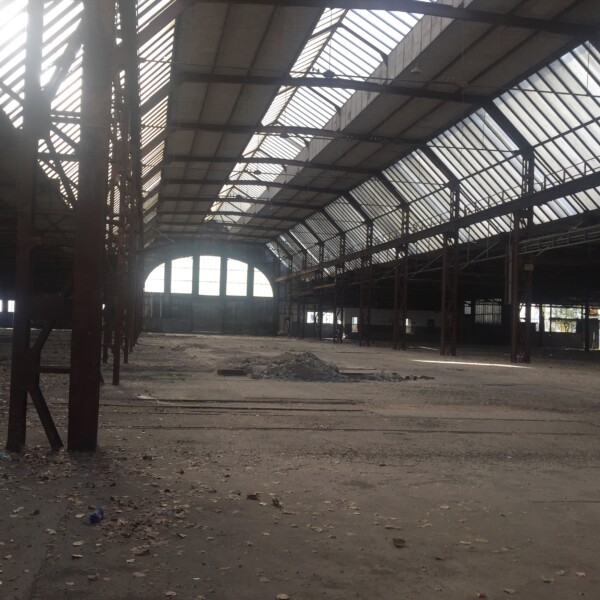 Innenansicht des Seilereigebäudes. Die Halle steht leer. Auf dem Boden sind Laub und in der Mitte ein Haufen Schutt zu sehen. An der Decke sind drei durchgängige Fensterreihen zu sehen.