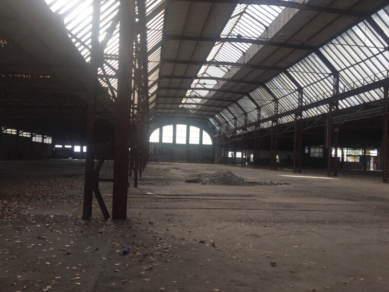 Innenansicht des Seilereigebäudes. Die Halle steht leer. Auf dem Boden sind Laub und in der Mitte ein Haufen Schutt zu sehen. An der Decke sind drei durchgängige Fensterreihen zu sehen.