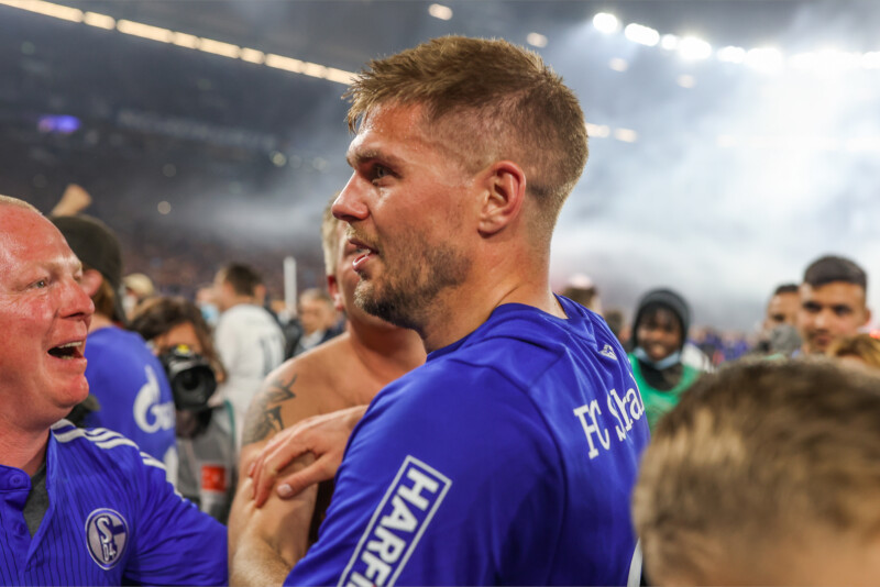 Simon Terodde steht verschwitzt im Schalke-Trikot in einer Menge jubelnder Menschen auf dem Spielfeld der Arena.