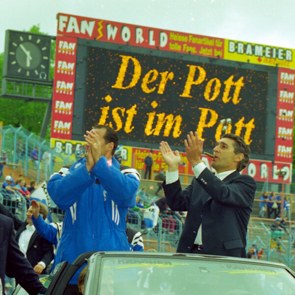 Huub Stevens und Rudi Assauer stehen in einem Cabrio. Sie lassen sich durchs Parkstadion fahren und feiern mit dem Publikum. Auf der Anzeigetafel im Hintergrund steht: "Der Pott ist im Pott".