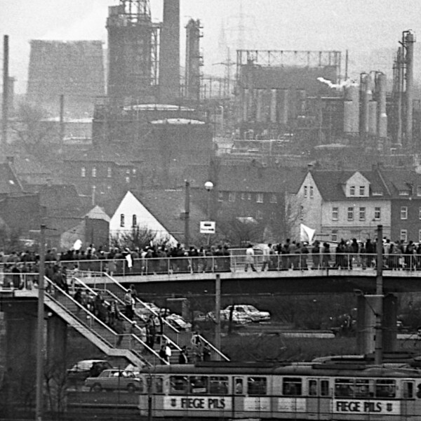 Menschen laufen über eine Fußgängerbrücke. Von ihr führen zwei Treppen herunter zu Straßenbahnhaltestellen. Im Hintergrund sind große Industrieanlagen zu erkennen.