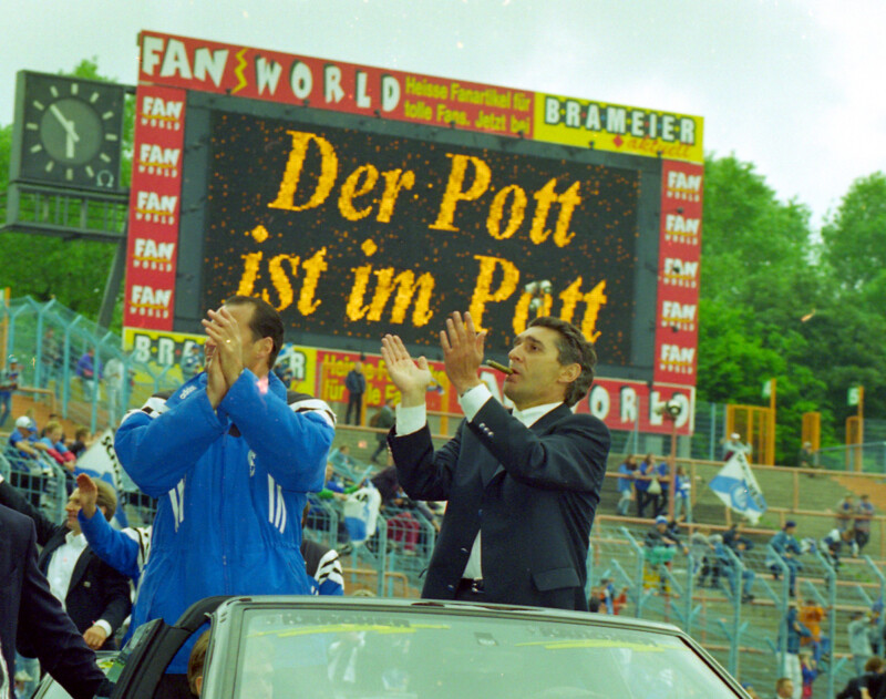 Huub Stevens und Rudi Assauer stehen in einem Cabrio. Sie lassen sich durchs Parkstadion fahren und feiern mit dem Publikum. Auf der Anzeigetafel im Hintergrund steht: "Der Pott ist im Pott".