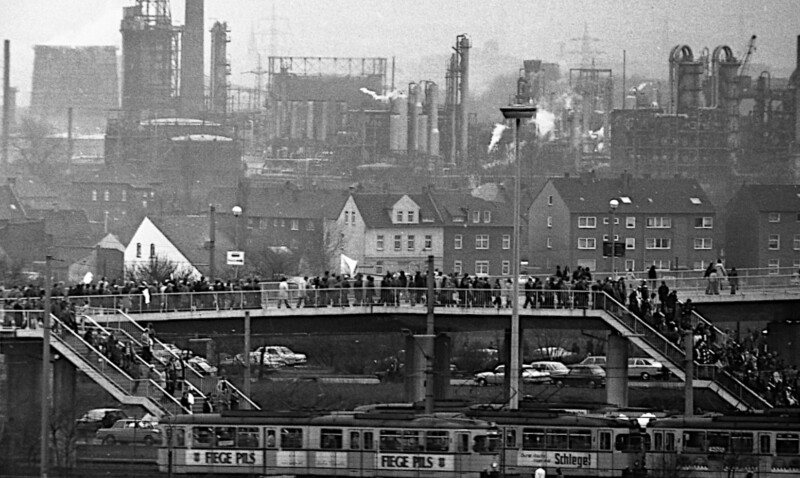 Menschen laufen über eine Fußgängerbrücke. Von ihr führen zwei Treppen herunter zu Straßenbahnhaltestellen. Im Hintergrund sind große Industrieanlagen zu erkennen.