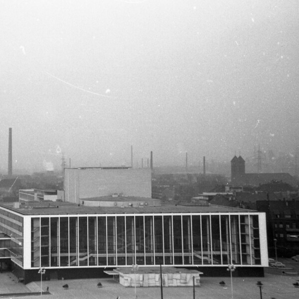 Vorderansicht des Musiktheaters im Revier in den 1950er-Jahren. Der moderne Bau verfügt über eine große Glasfassade. Dahinter erhebt sich die Schalker Industriekulisse mit Zeche Consolidation 1/6. Rechts ist die St.-Joseph-Kirche zu sehen.