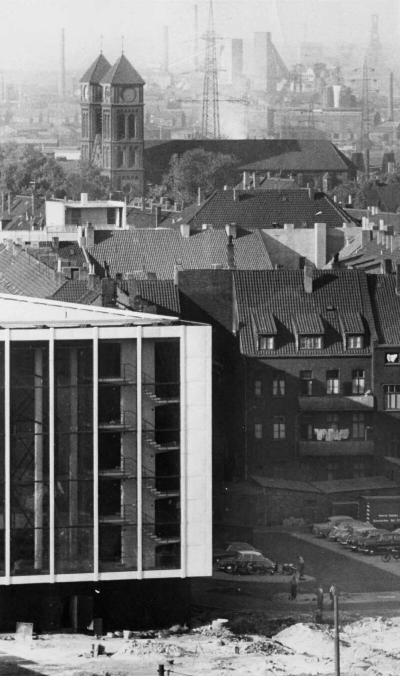 Detailansicht der rechten Ecke der Glasfassade des Musiktheaters im Revier. Im Hintergrund stehen die St.-Joseph-Kirche und Teile der Schalker Industrie.