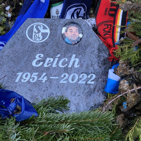 Der Grabstein von Erich Wehner auf dem Schalker FanFeld, eingerahmt von einem Schalke-Schal und einem Schal des 1. FC Nürnberg. Zwischen den Schals steht eine Bierflasche der Marke Veltins.