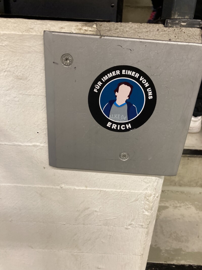 Auf eine Mauer ist ein Sticker mit einer stilisierten Darstellung von Erich Wehner geklebt. Dazu steht auf dem Sticker: "Für immer einer von uns - Erich".