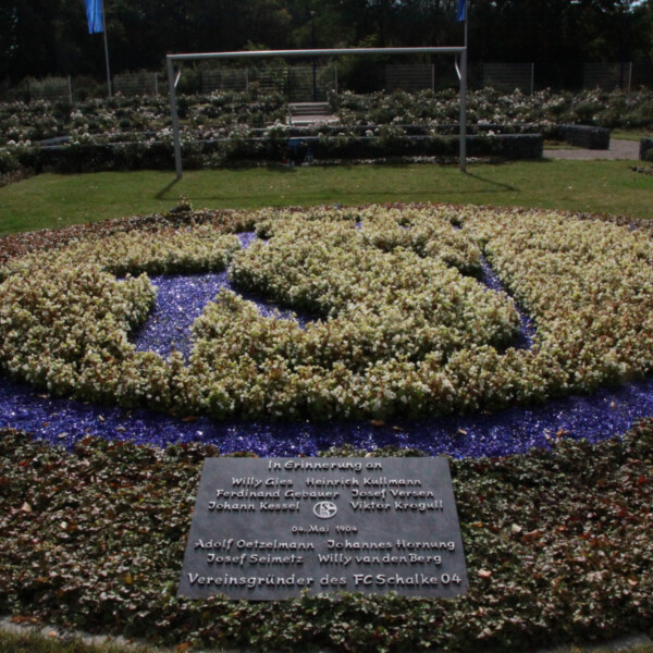 Ein Beet aus blauen und weißen Blumen, die das Wappen des FC Schalke 04 formen. Davor liegt ein Gedenkstein, der an die Menschen erinnert, die 1904 den Verein gründeten.