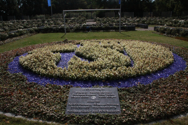 Ein Beet aus blauen und weißen Blumen, die das Wappen des FC Schalke 04 formen. Davor liegt ein Gedenkstein, der an die Menschen erinnert, die 1904 den Verein gründeten.