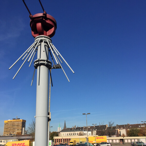 Eine Protegohaube steht hinter einer kleinen Betonmauer. Die Protegohaube besteht aus einem grauen Rohr, das etwa so dick ist wie eine Fallrohr. Da drauf sitzt ein roter Verschluss. Dahinter liegt ein Parkplatz.