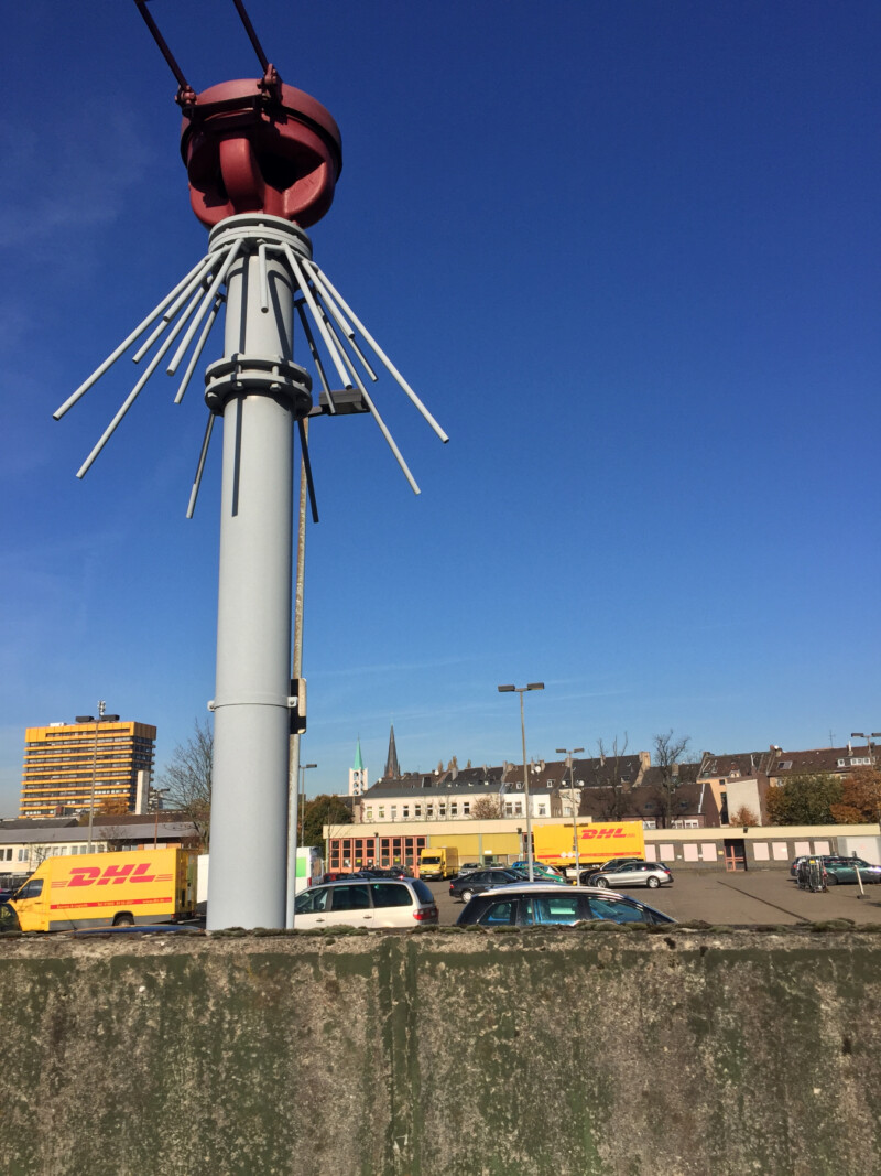 Eine Protegohaube steht hinter einer kleinen Betonmauer. Die Protegohaube besteht aus einem grauen Rohr, das etwa so dick ist wie eine Fallrohr. Da drauf sitzt ein roter Verschluss. Dahinter liegt ein Parkplatz.