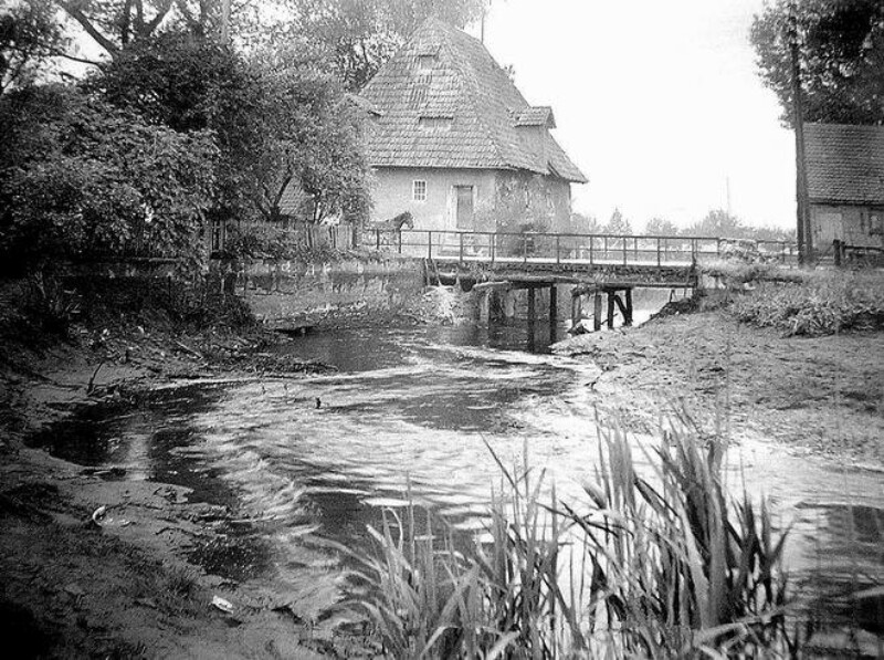 Unter einer Brücke, die von einem Pferdegespann überquert wird, fließt ein Fluss. An der Brücke steht ein Haus, ansonsten ist die Natur unberührt.