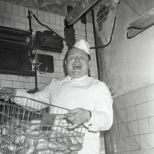 Der junge Karlheinz "Charly" Neumann hält einen Drahtkorb mit Brötchen hoch. Er trägt die typische Arbeitskleidung eines Bäckers.