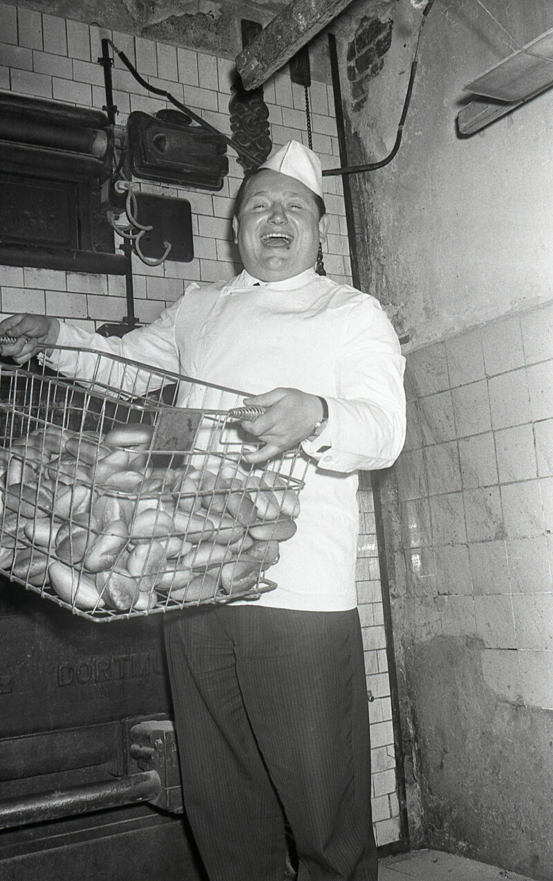 Der junge Karlheinz "Charly" Neumann hält einen Drahtkorb mit Brötchen hoch. Er trägt die typische Arbeitskleidung eines Bäckers.